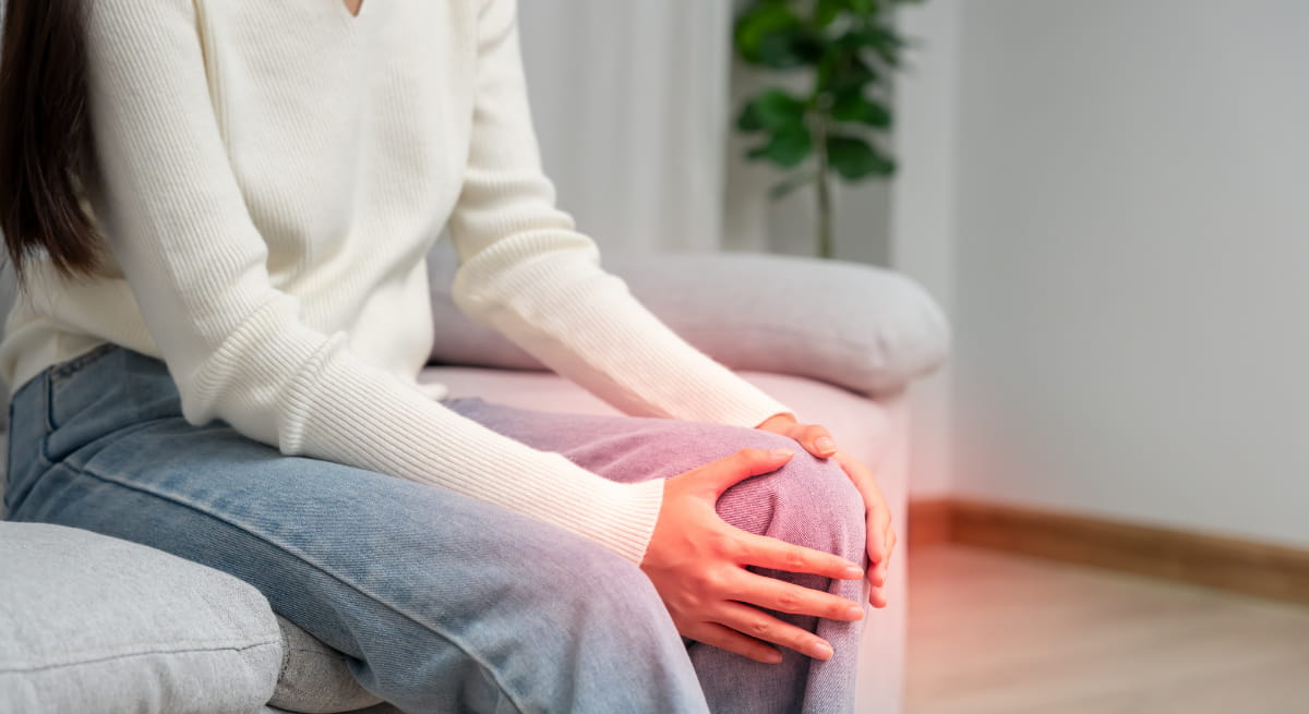 Person sitting on a sofa, fully dressed, resting one hand on a sore knee